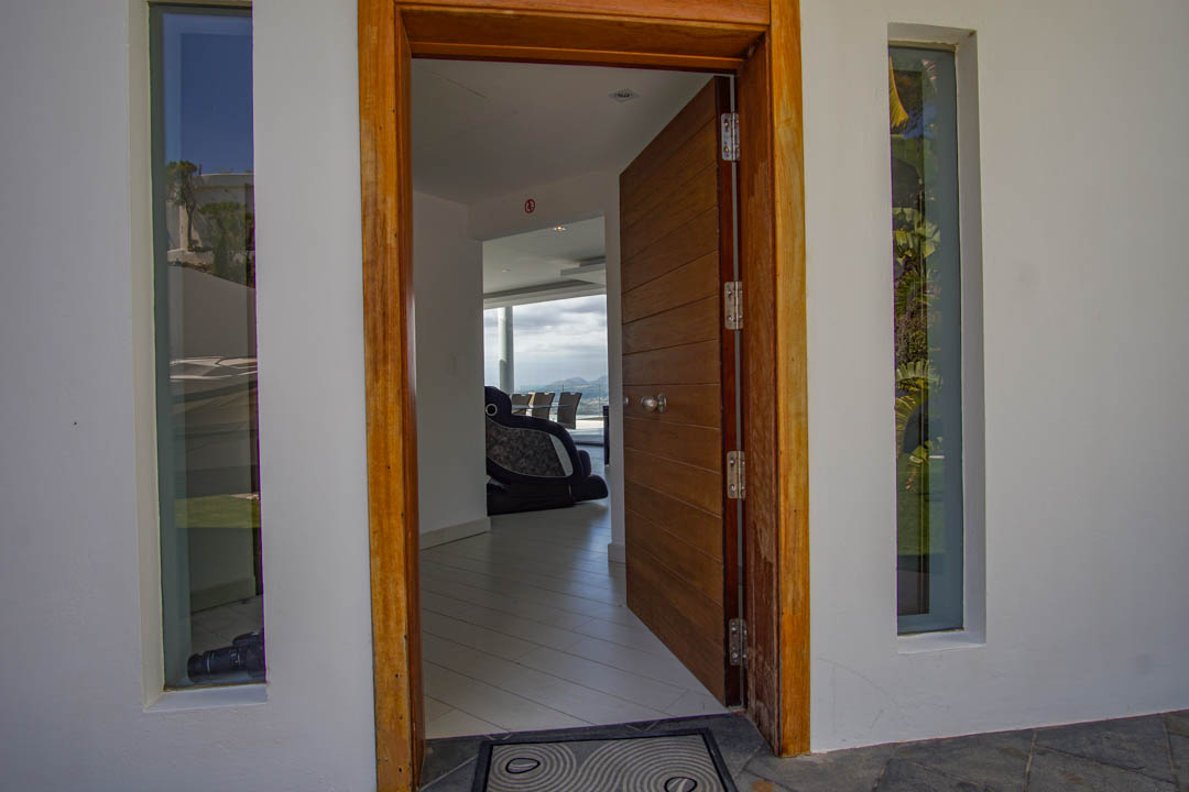 L'entrée principale ouverte révèle une porte et un cadre en bois à panneaux, flanqués de fenêtres verticales, offrant un aperçu de l'intérieur moderne et des vues sur la montagne.