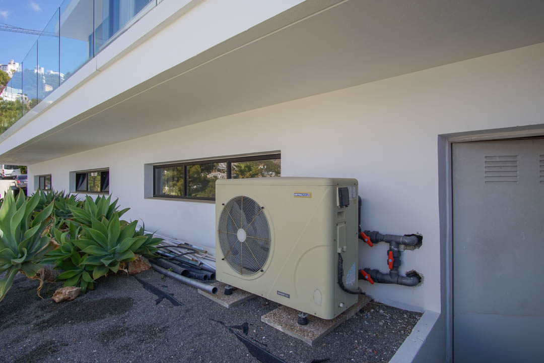 L'espace technique extérieur de la villa présente une pompe à chaleur inverter AstralPool, des fenêtres horizontales et une porte de service. Des agaves complètent la façade moderne.