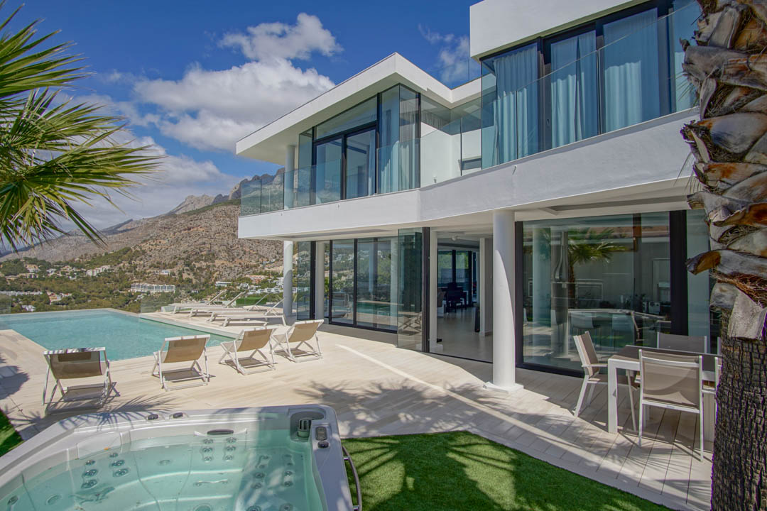 L'extérieur de la villa blanche moderne révèle une piscine à débordement, un jacuzzi séparé et une vaste terrasse avec transats, encadrée par des vues montagneuses sous un ciel clair.