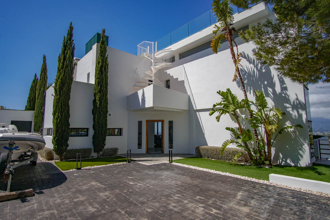 L'extérieur de la villa moderne présente une allée pavée, des cyprès élancés et un escalier en colimaçon blanc menant à la terrasse supérieure avec balustrade en verre.