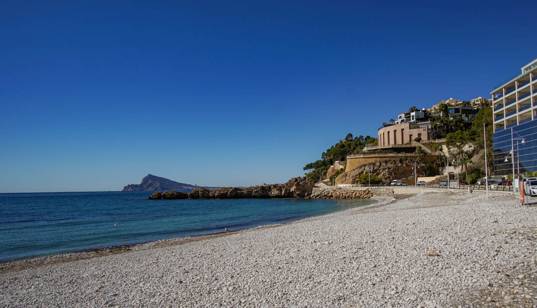 Plage de galets avec des eaux turquoise claires et une côte rocheuse. Des bâtiments modernes aux façades réfléchissantes et une structure arrondie bordent la colline adjacente, sous un ciel bleu.