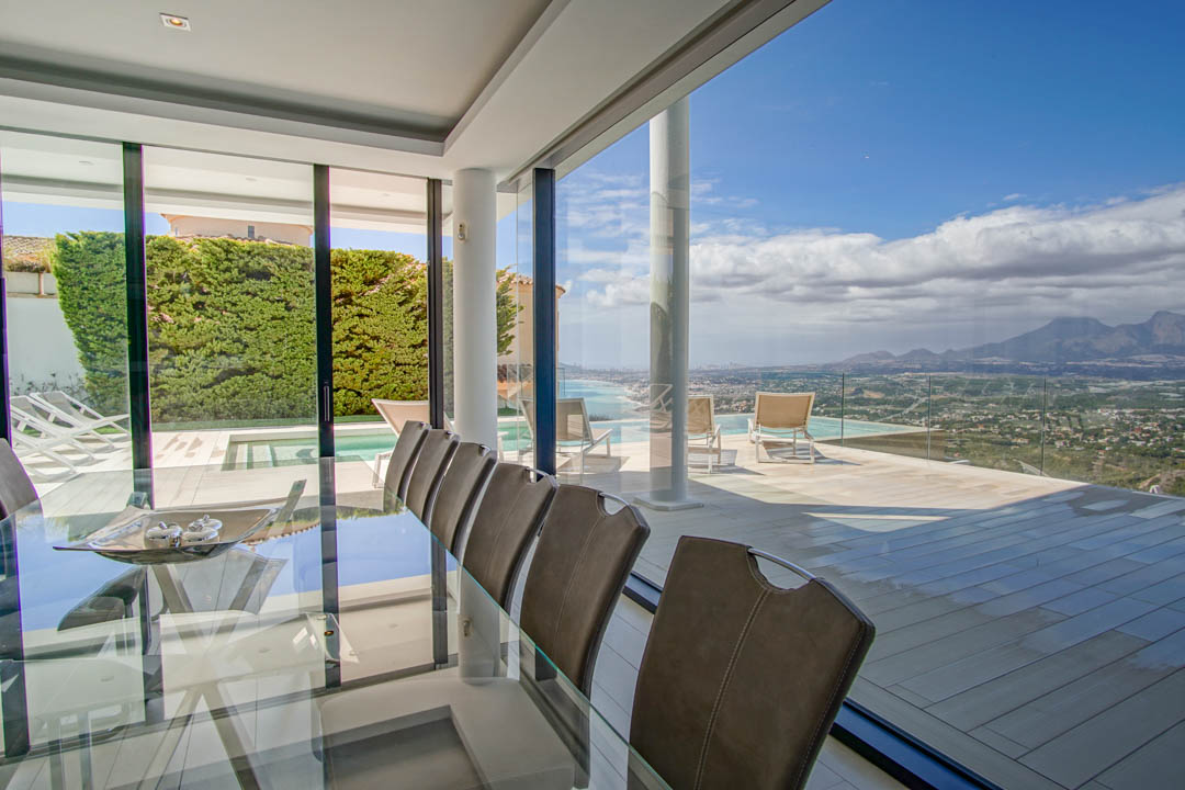 Salle à manger intérieure élégante avec table en verre et chaises, s'ouvrant sur une vaste terrasse avec piscine à débordement et vues panoramiques sur la mer et les montagnes.