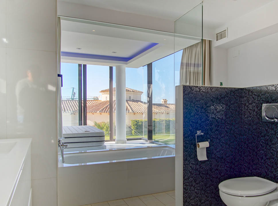 Salle de bain contemporaine avec baignoire intégrée et fenêtres du sol au plafond donnant sur un toit de tuiles et un jardin. Comprend un éclairage d'accentuation bleu et une cloison à motif sombre.