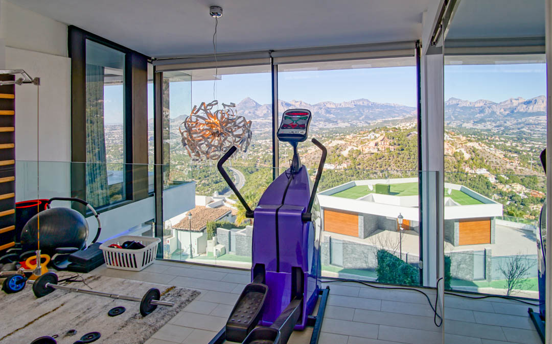 Salle de sport avec baies vitrées offrant une vue panoramique sur les montagnes et la vallée. Équipée d'un vélo elliptique et de poids, l'espace intègre une balustrade en verre.