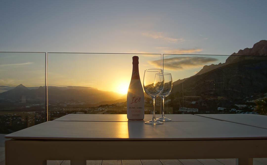 Table d'extérieur sur une terrasse avec balustrade en verre, présentant une bouteille de cava rosé et deux verres. Coucher de soleil sur les montagnes et la vallée, reflets chaleureux.