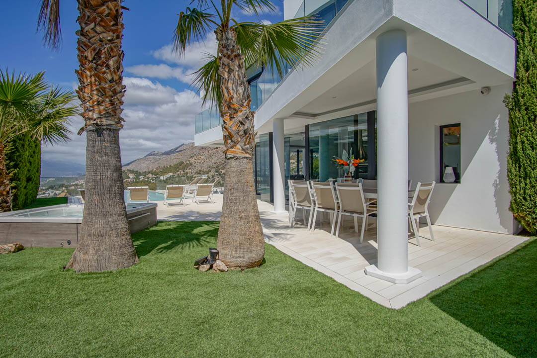 Terrasse couverte pour les repas en plein air avec mobilier moderne, bordée d'une pelouse artificielle et de palmiers. Un jacuzzi et la piscine offrent des vues sur les montagnes et la mer.