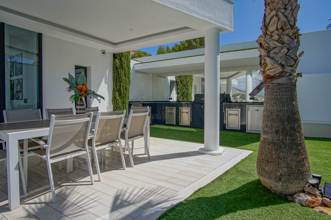 Terrasse extérieure avec salle à manger sur un sol carrelé clair et gazon artificiel. Une cuisine extérieure couverte avec barbecue et appareils en acier inoxydable est visible, près d'un palmier.