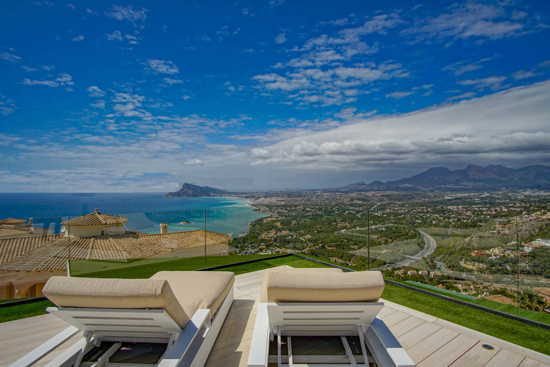 Terrasse sur le toit avec deux chaises longues blanches sur une terrasse en bois clair et gazon artificiel, offrant une vue panoramique sur la mer Méditerranée, la côte d'Altea et les montagnes.