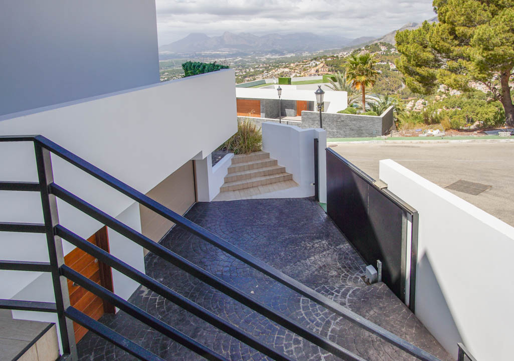 Un escalier extérieur moderne avec garde-corps noirs descend vers une allée texturée gris foncé, dotée d'un portail automatique, d'une entrée de garage et de vues panoramiques sur les montagnes.
