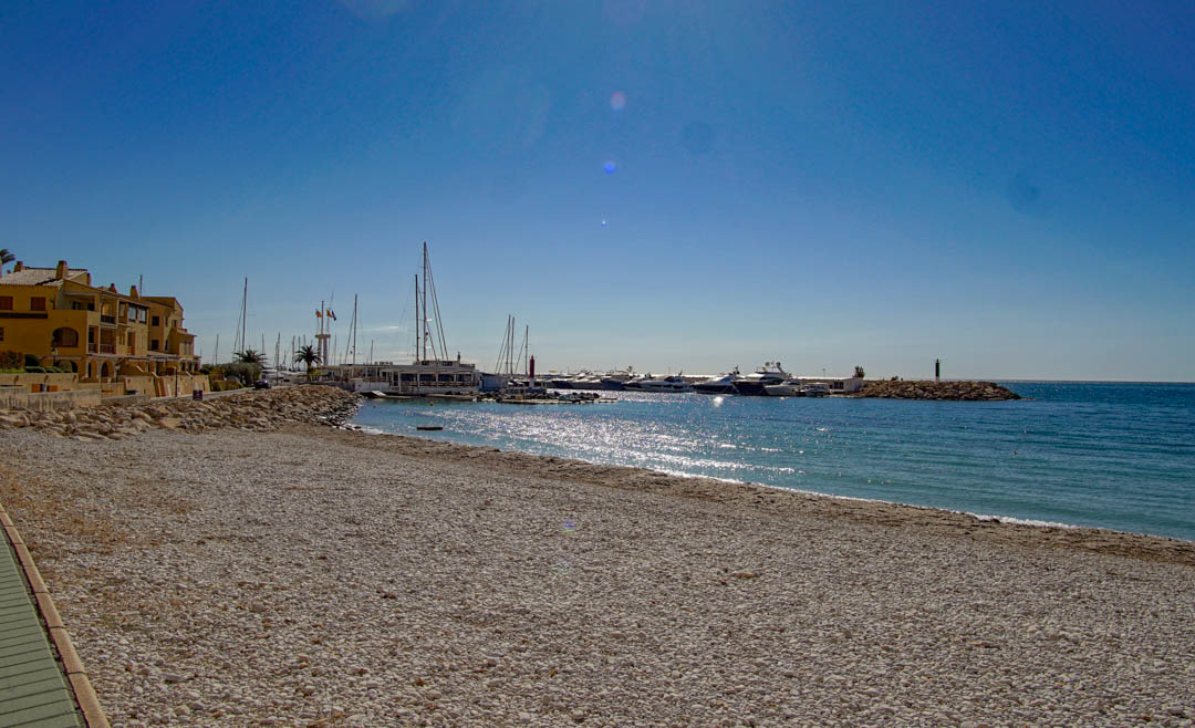 Une large plage de galets s'étend vers une marina remplie de yachts, bordée de bâtiments côtiers traditionnels sous un ciel bleu clair.