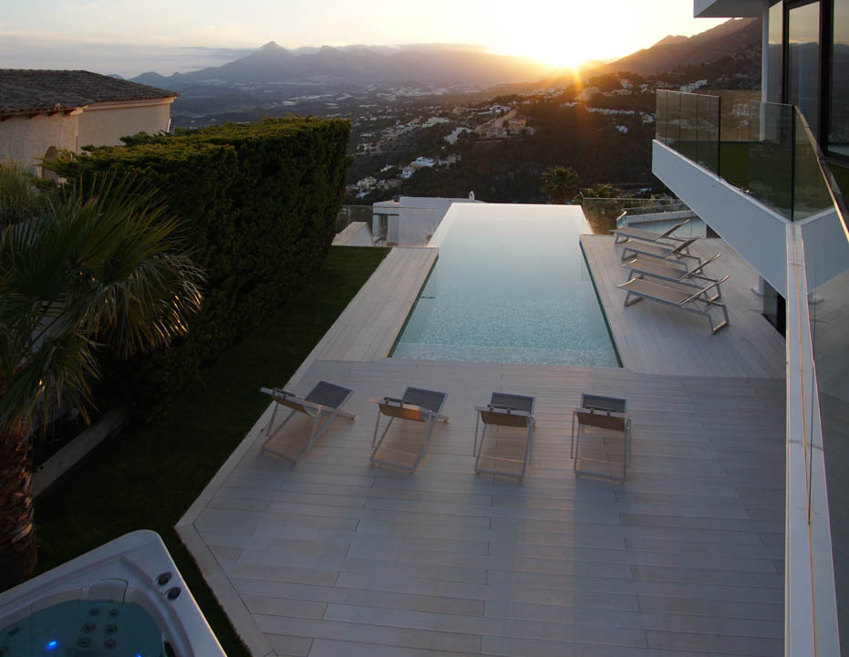 Vaste piscine à débordement avec terrasse claire, chaises longues et jacuzzi séparé, offrant une vue panoramique sur un coucher de soleil montagneux.