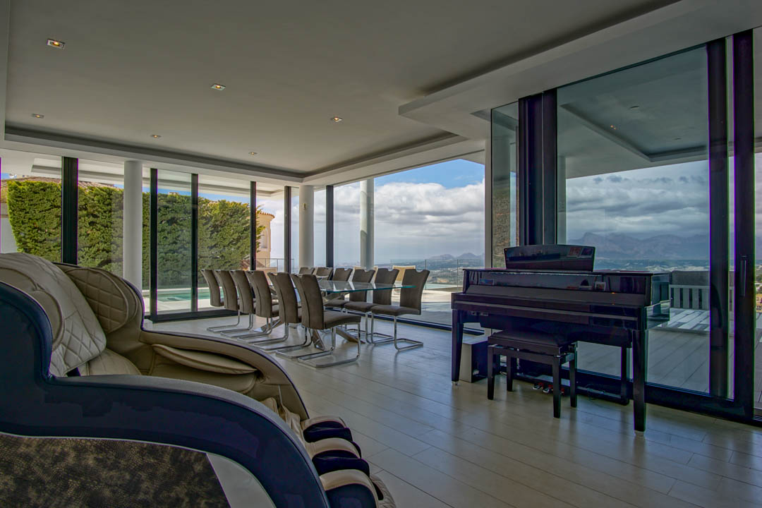 Vaste salon-salle à manger avec table en verre pour dix, piano à queue et fauteuil de massage. Grandes baies vitrées offrant des vues panoramiques sur les montagnes et le paysage.