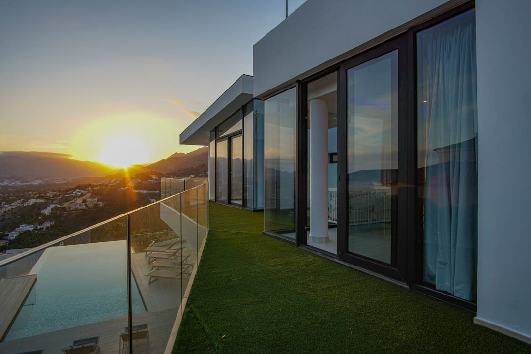 Vaste terrasse avec piscine à débordement, pont en bois et balustrade en verre, offrant des vues panoramiques sur le coucher de soleil montagneux.