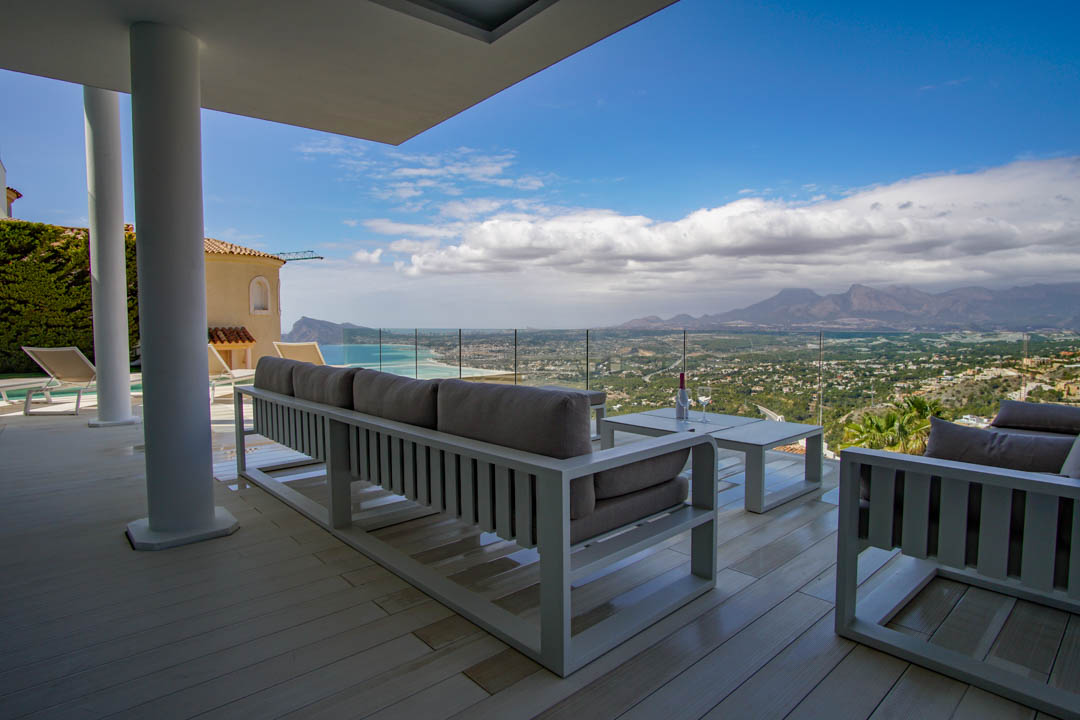Vaste terrasse couverte avec plancher clair et mobilier lounge blanc moderne, offrant une vue panoramique sur la mer, la côte et les montagnes via une balustrade vitrée.