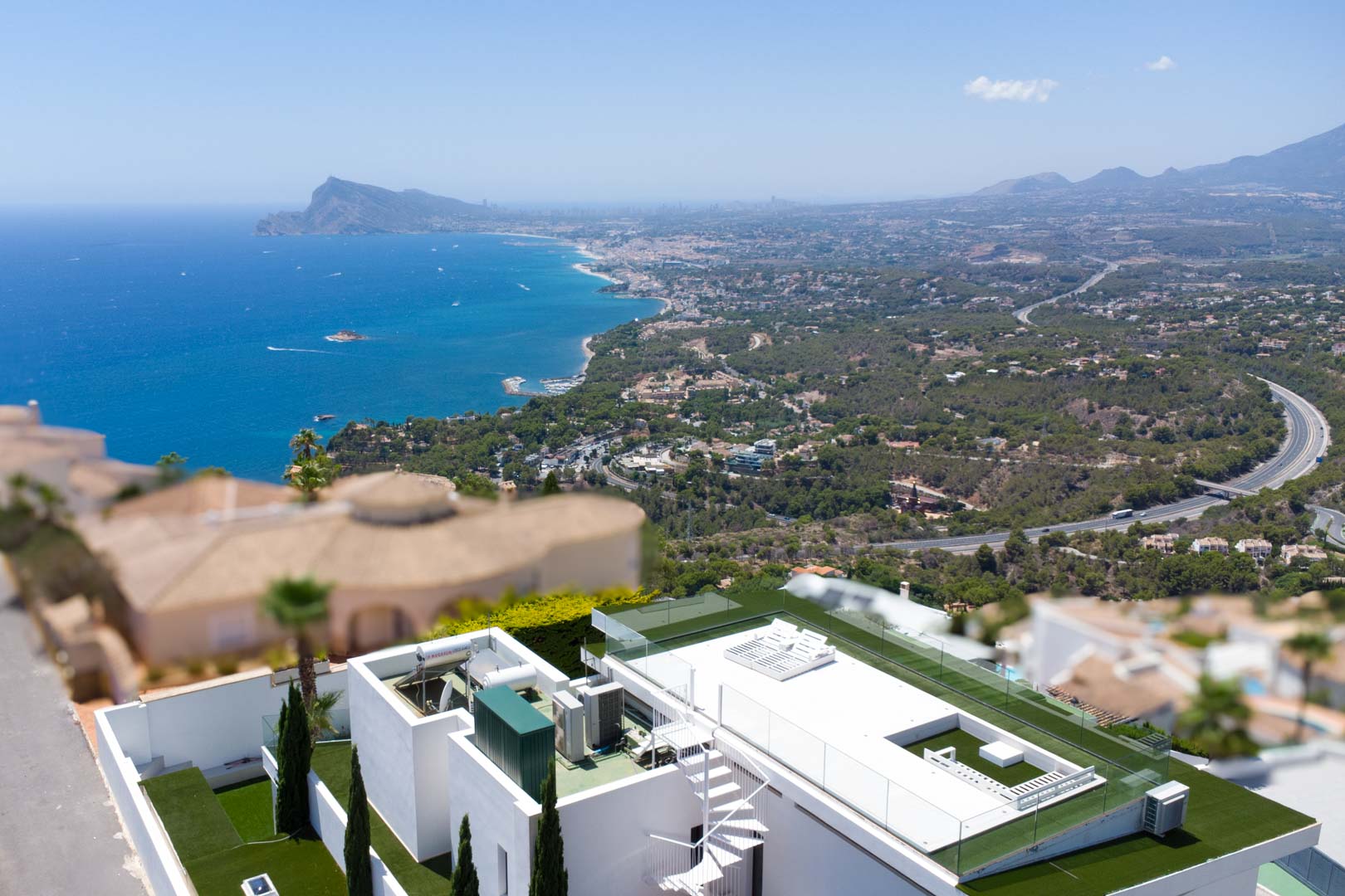 Vue aérienne de la terrasse sur le toit de la villa, dotée d'un escalier en colimaçon blanc, de gazon artificiel, de garde-corps en verre et de vues panoramiques sur la mer et la côte.