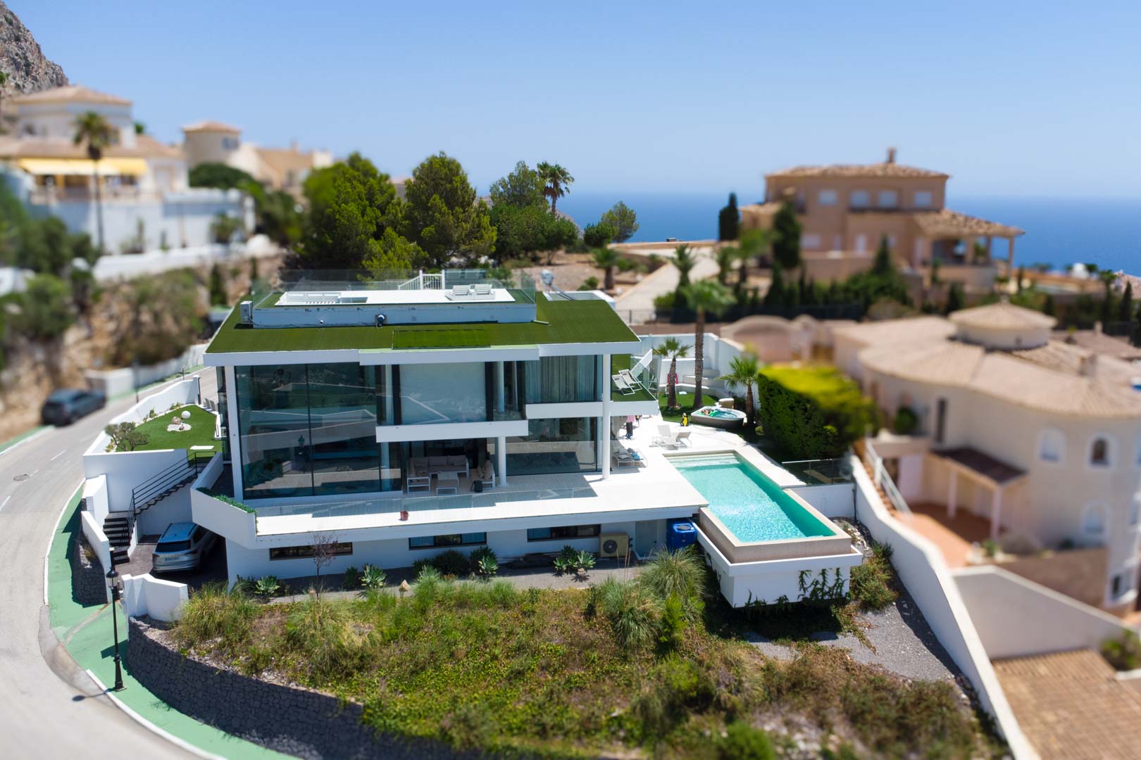 Vue aérienne de la villa moderne blanche au design épuré avec toit végétalisé, piscine à débordement et jacuzzi séparé, offrant une vue sur la mer.