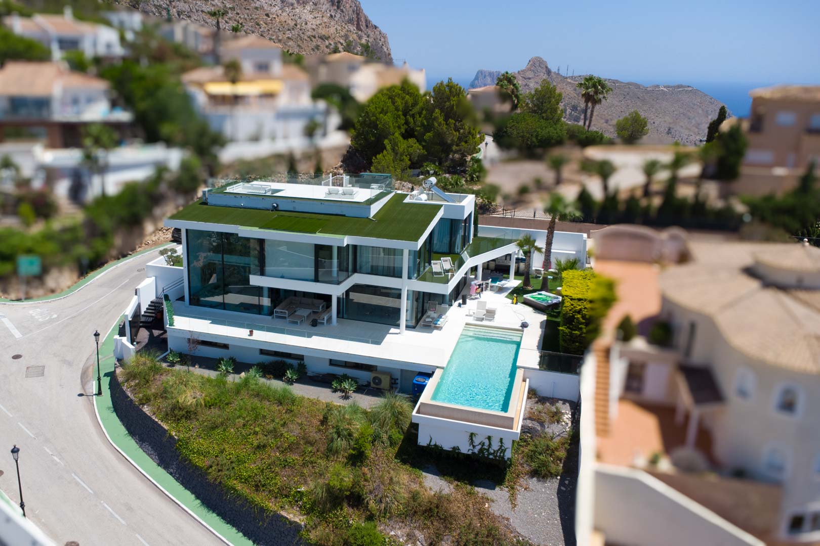 Vue aérienne d'une villa blanche contemporaine avec piscine à débordement, façades vitrées, toit végétalisé et jacuzzi, offrant des vues sur la montagne et la mer.