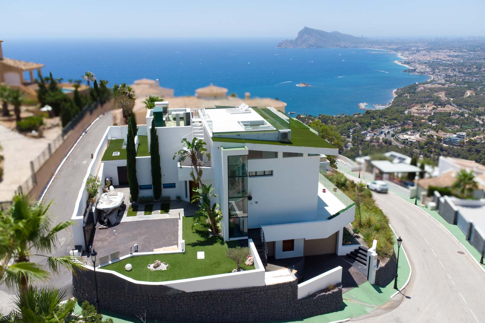 Vue aérienne d'une villa blanche moderne à plusieurs niveaux, avec toits plats végétalisés et ascenseur extérieur en verre. Elle dispose d'une allée privée et de vues panoramiques sur la mer et la côte.