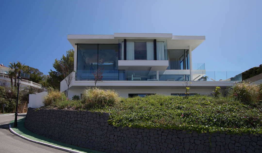 Vue extérieure d'une villa moderne à trois niveaux avec de vastes façades vitrées et balustrades en verre, surplombant un mur de pierre et la végétation le long d'une route courbée.