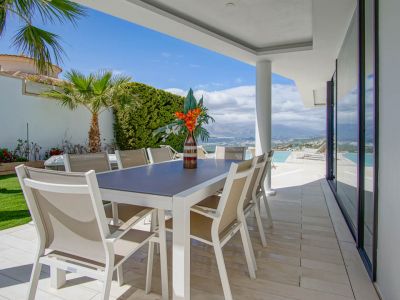 Cette terrasse repas extérieure couverte dispose d'une table blanche moderne et de huit chaises beiges sur carrelage clair. Elle offre des vues étendues sur les montagnes et la mer, à côté de la piscine à débordement.