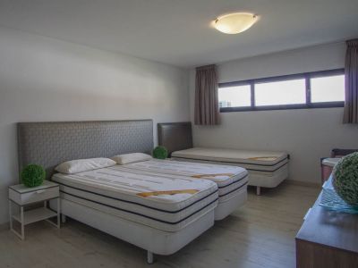 Chambre à coucher avec deux lits simples combinés, têtes de lit grises texturées, un troisième lit simple à l'arrière, parquet clair et fenêtres horizontales.