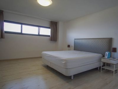 Chambre à coucher avec sol aspect bois clair et murs blancs. Comprend un lit avec tête de lit texturée, une table de chevet et une fenêtre horizontale encadrée de noir.