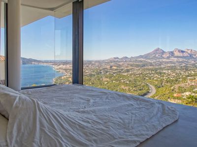 Chambre avec baies vitrées du sol au plafond, encadrant des vues panoramiques sur la mer Méditerranée, une ville côtière et les montagnes lointaines. Un lit bas avec linge blanc fait face à la vue.