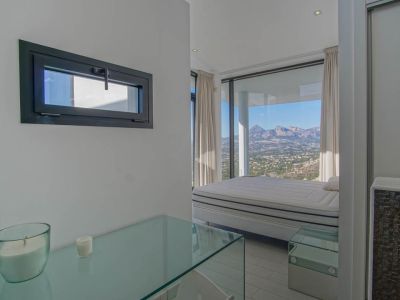 Chambre lumineuse avec un lit placé devant de grandes baies vitrées, offrant une vue panoramique sur les montagnes et la vallée. Une console en verre et une fenêtre horizontale sont visibles.