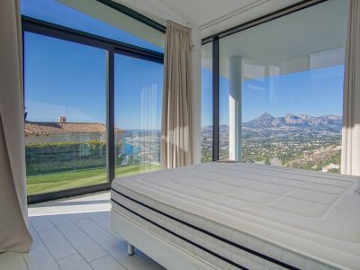 Chambre moderne avec grand lit et vastes fenêtres. Elle offre des vues panoramiques sur la mer lointaine, une pelouse verte et des montagnes escarpées sous un ciel clair.