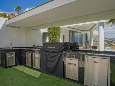 Cuisine extérieure en L avec plans de travail en granit noir, double évier, barbecue couvert et appareils en acier inoxydable, reflétant la vue sur la piscine.