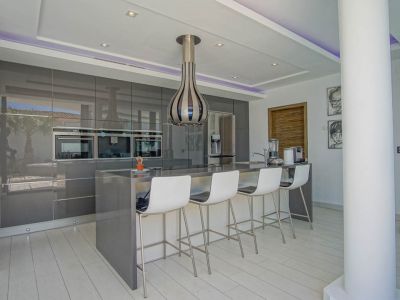 Cuisine moderne avec des armoires grises laquées, fours encastrés et un îlot central servant de bar pour quatre personnes. Éclairage LED intégré et reflets extérieurs.