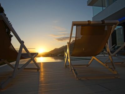 Deux chaises longues modernes sur une terrasse claire surplombent une piscine à débordement, reflétant un coucher de soleil doré sur les montagnes.