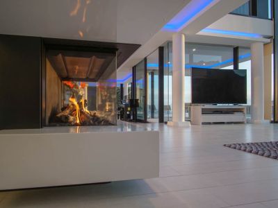 Espace de vie moderne avec cheminée à gaz vitrée et base blanche. De grandes fenêtres et un éclairage d'accentuation bleu complètent le design.