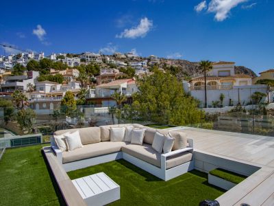 Espace salon sur le toit avec canapé en L sur gazon artificiel, table basse blanche et garde-corps en verre. Offre des vues dégagées sur la colline en terrasses et ses résidences.