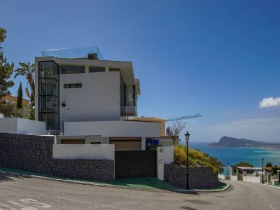 Extérieur d'une villa moderne blanche avec un ascenseur en verre et une balustrade de terrasse sur le toit. Murs de soutènement en pierre et portail automatique, avec vue panoramique sur la mer.