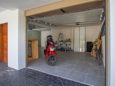 Grand garage avec carrelage gris, porte sectionnelle partiellement ouverte et porte d'entrée en bois distinctive à gauche. Une trottinette rouge et un vélo sont visibles.