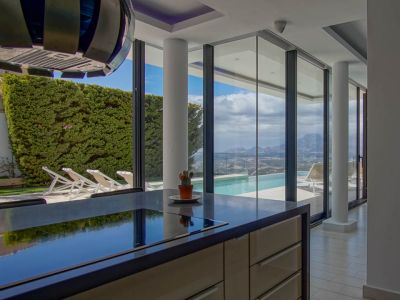 Îlot de cuisine moderne avec plan de travail sombre et plaque de cuisson intégrée. Les baies vitrées offrent une vue sur la piscine à débordement, la vallée et les montagnes.