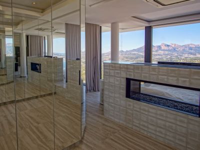 Intérieur contemporain avec cheminée à gaz transparente sur lit de galets, intégrée dans un mur texturé. Des panneaux miroir reflètent la pièce et les vues imprenables sur les montagnes et la mer.