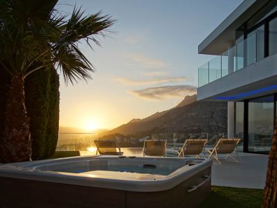 Jacuzzi extérieur avec finition bois, sur pelouse verte, devant une villa moderne avec balcons vitrés. Vue montagne et mer au coucher du soleil.