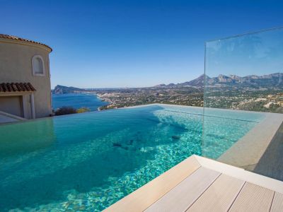 La piscine à débordement avec son eau claire, sa terrasse claire et sa barrière en verre, offre une vue panoramique sur la mer, la côte et les montagnes lointaines.