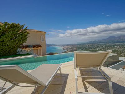 La terrasse extérieure comprend une piscine à débordement offrant une vue sur le littoral et les montagnes lointaines. Deux chaises longues sur un pont clair, bordées d'une balustrade en verre et d'une façade de villa.