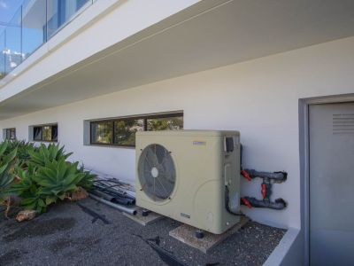 L'espace technique extérieur de la villa présente une pompe à chaleur inverter AstralPool, des fenêtres horizontales et une porte de service. Des agaves complètent la façade moderne.