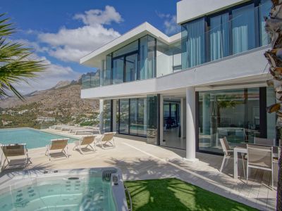 L'extérieur de la villa blanche moderne révèle une piscine à débordement, un jacuzzi séparé et une vaste terrasse avec transats, encadrée par des vues montagneuses sous un ciel clair.