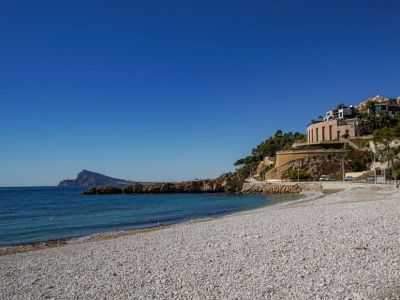 Plage de galets avec des eaux turquoise claires et une côte rocheuse. Des bâtiments modernes aux façades réfléchissantes et une structure arrondie bordent la colline adjacente, sous un ciel bleu.