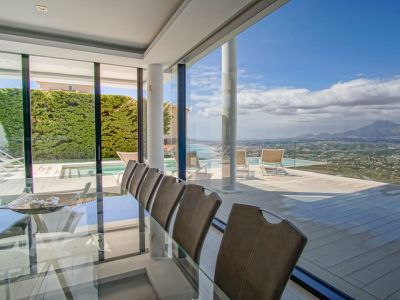 Salle à manger intérieure élégante avec table en verre et chaises, s'ouvrant sur une vaste terrasse avec piscine à débordement et vues panoramiques sur la mer et les montagnes.