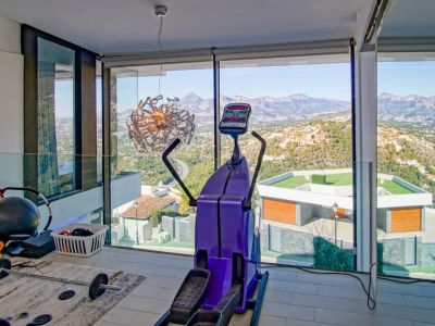 Salle de sport avec baies vitrées offrant une vue panoramique sur les montagnes et la vallée. Équipée d'un vélo elliptique et de poids, l'espace intègre une balustrade en verre.