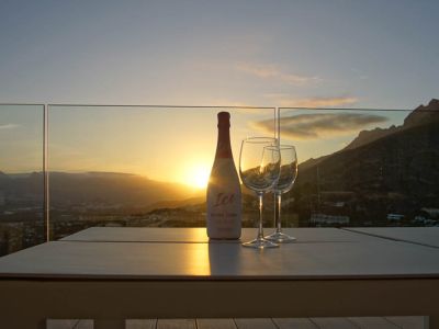 Table d'extérieur sur une terrasse avec balustrade en verre, présentant une bouteille de cava rosé et deux verres. Coucher de soleil sur les montagnes et la vallée, reflets chaleureux.