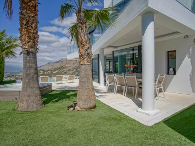 Terrasse couverte pour les repas en plein air avec mobilier moderne, bordée d'une pelouse artificielle et de palmiers. Un jacuzzi et la piscine offrent des vues sur les montagnes et la mer.