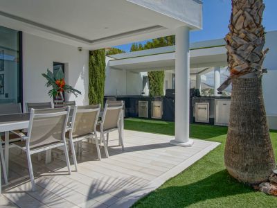 Terrasse extérieure avec salle à manger sur un sol carrelé clair et gazon artificiel. Une cuisine extérieure couverte avec barbecue et appareils en acier inoxydable est visible, près d'un palmier.