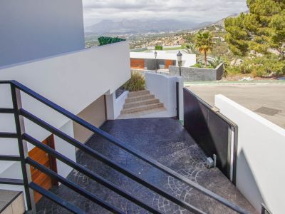 Un escalier extérieur moderne avec garde-corps noirs descend vers une allée texturée gris foncé, dotée d'un portail automatique, d'une entrée de garage et de vues panoramiques sur les montagnes.
